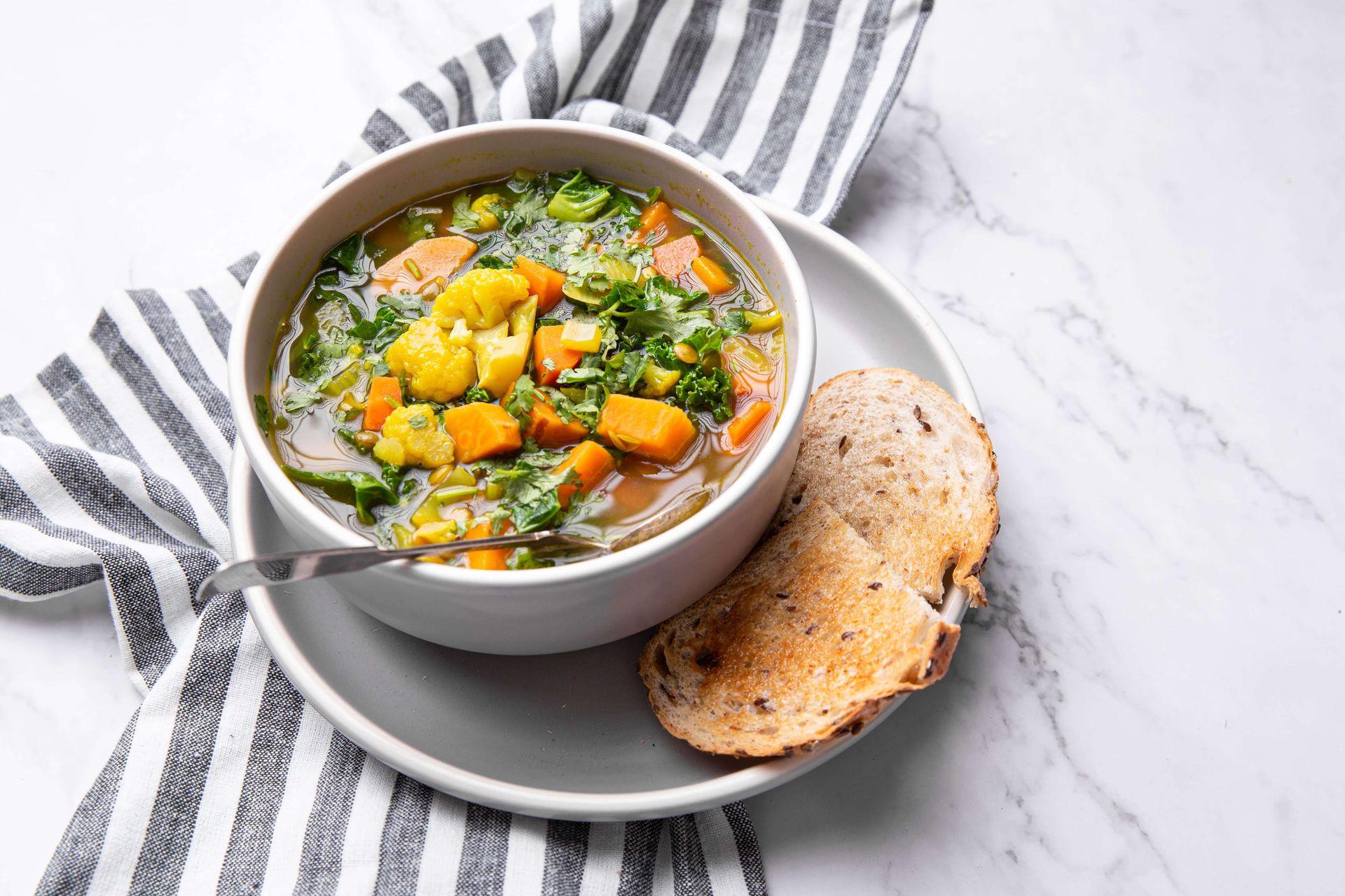 Bowl of vegetable soup with bread on a striped cloth and marble surface.